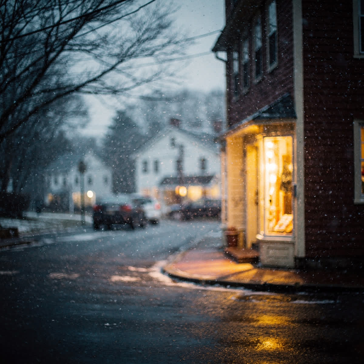 Snow falling at dusk, warm light from a shop window